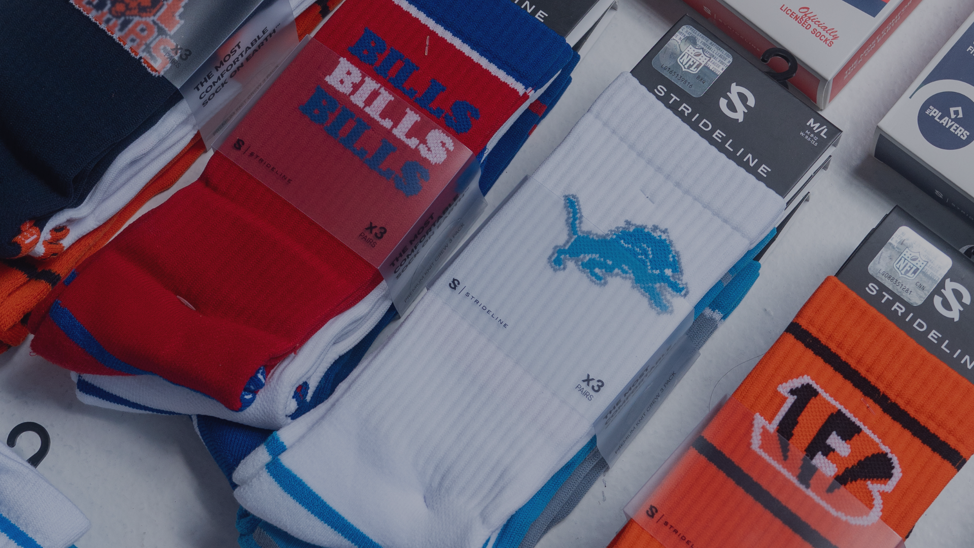 A close-up, angled view of multiple packaged pairs of athletic crew socks spread out on a white surface. Visible socks include a red pair with blue trim and 'BILLS BILLS BILLS' text, a white pair with a blue lion logo and 'STRIDELINE' packaging, an orange pair with black stripes and a white '1F3' graphic, and other partially visible socks in navy, white, and orange.