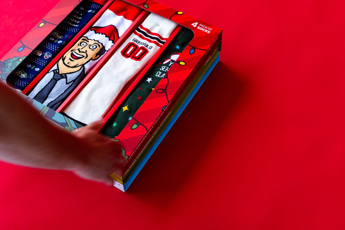 Colorful holiday-themed socks in a gift box with a hand reaching for them, set against a red background.